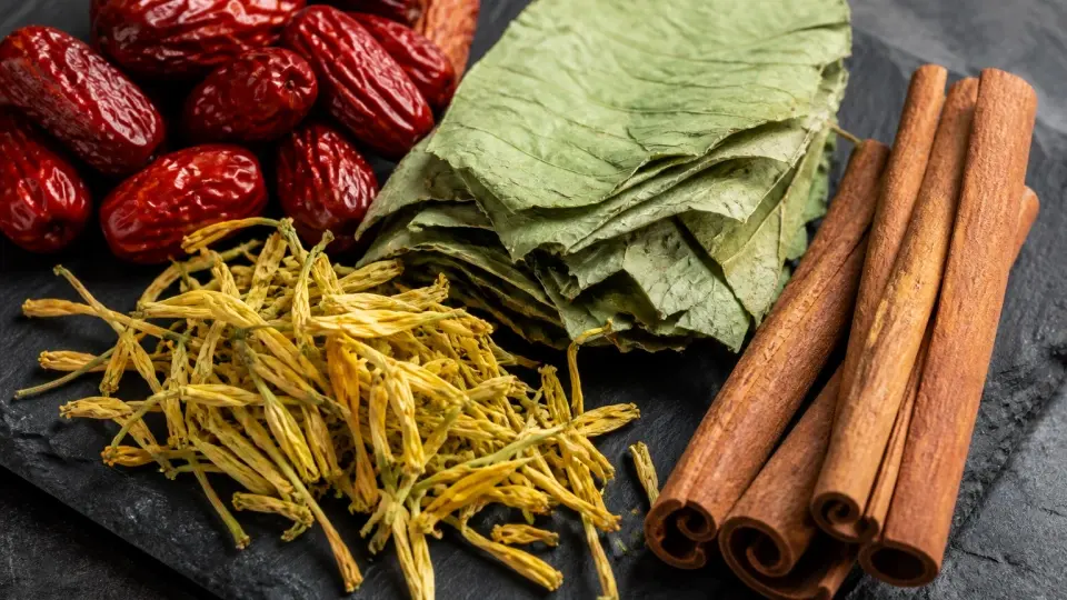 A detailed close-up shot of dried red dates, green lotus leaves, golden honeysuckle, and textured cinnamon bark